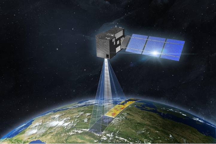The image is an artist’s impression of a CO2M satellite in space. The satellite has a rectangular body with solar panels extending from one side. It is positioned above Earth, with a visible beam projecting downward, indicating the satellite's monitoring capabilities. The Earth's surface below shows a detailed section of land with varying colours, likely representing data collected by the satellite. The background features the dark expanse of space dotted with stars. This satellite image is credited to OHB